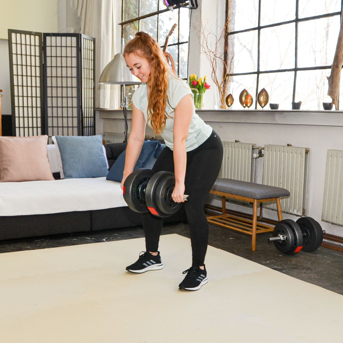 Eine Person hebt Gewichte auf einer Fitnessmatte. Die Person steht aufrecht und hält zwei Hanteln.