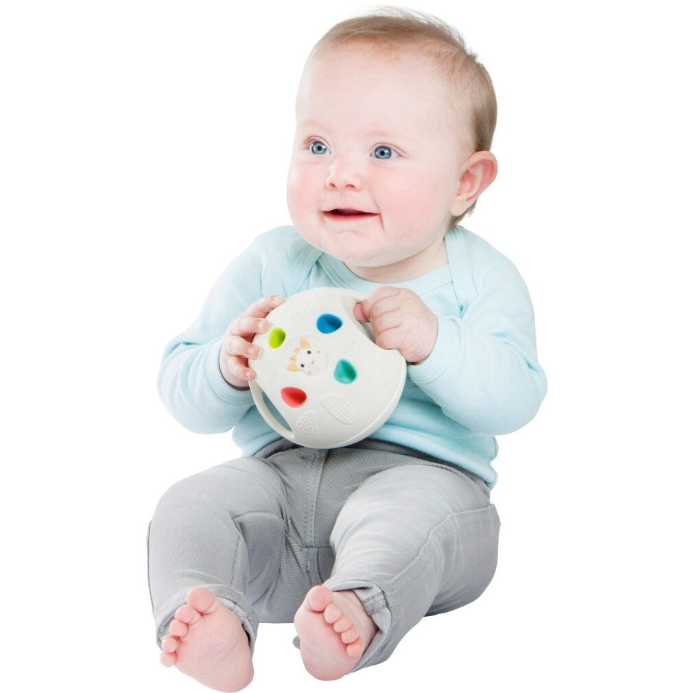 Baby hält weißen Ball mit farbigen Elementen und Giraffen-Gesicht. Baby sitzt. Helle Kleidung.