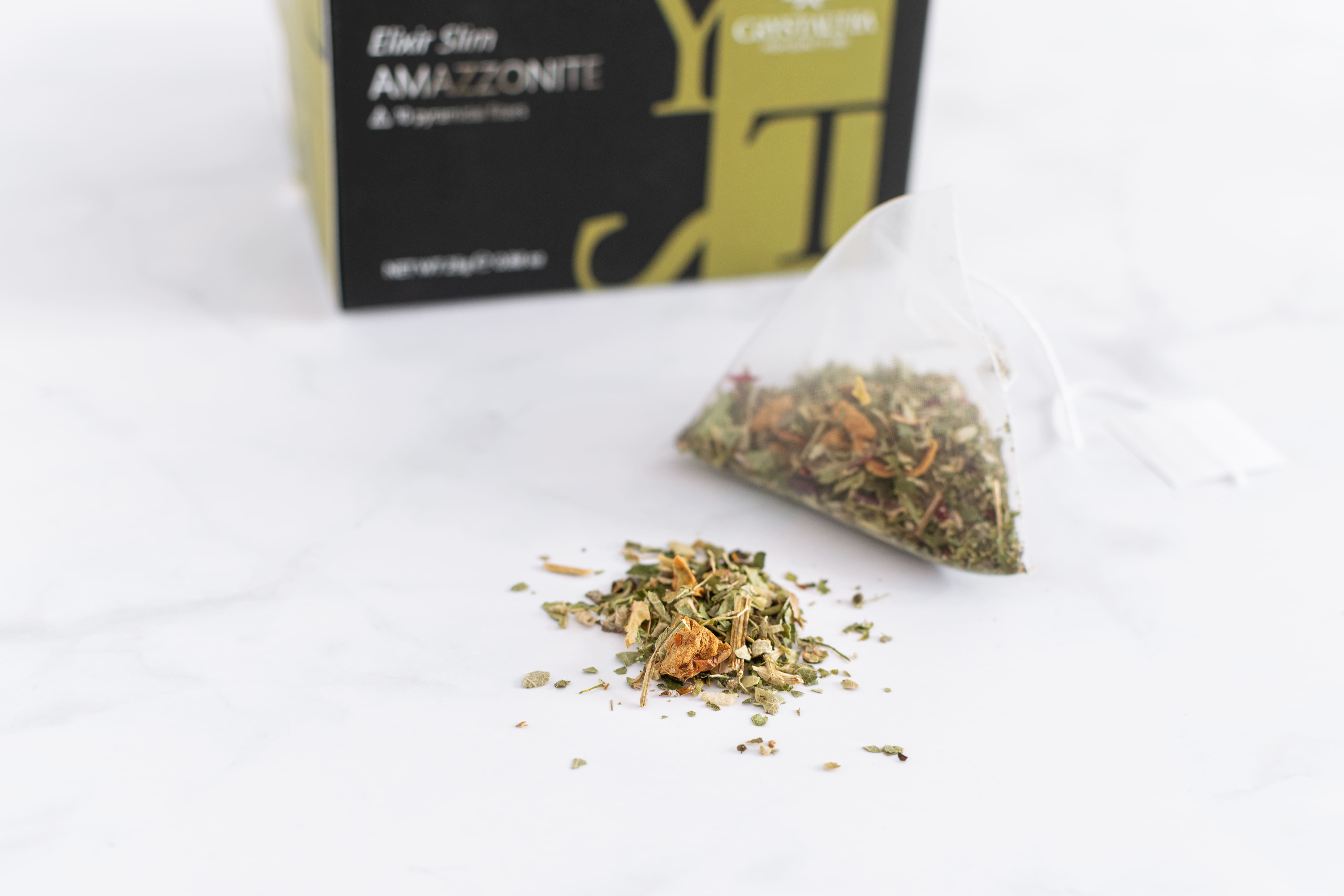 Teebeutel mit Kräutern und lose Kräuter daneben. Im Hintergrund eine Schachtel mit Produktnamen und Logo.