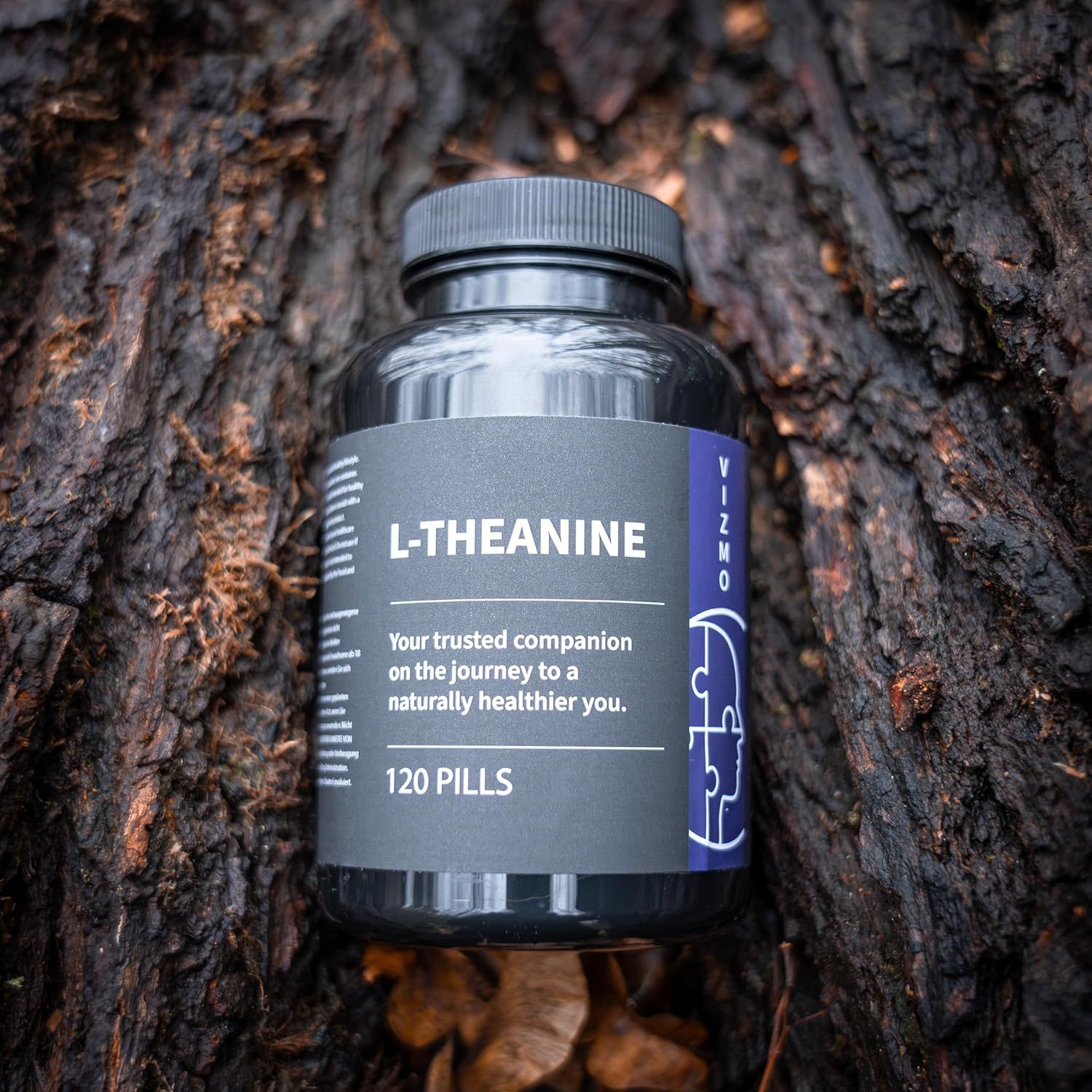 Schwarze Flasche L-Theanin-Kapseln auf Baumrinde. Aufschrift: L-Theanin, 120 Pillen. Marke: Vizmo. Lila Etikett mit Kopf-Puzzle-Grafik.