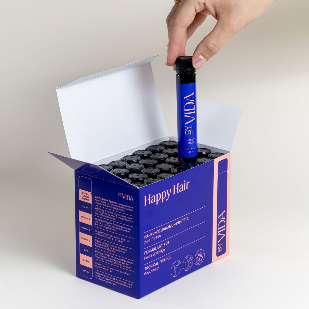 Geöffnete blaue Box mit "Happy Hair BYVIDA" Trinkampullen. Eine Hand nimmt eine Ampulle heraus.