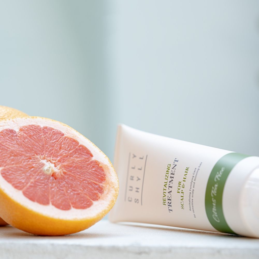 Beige Tube mit grünem Streifen. Text: CURLY SHYLL, Revitalizing Treatment for Scalp & Hair. Daneben eine Grapefruit.