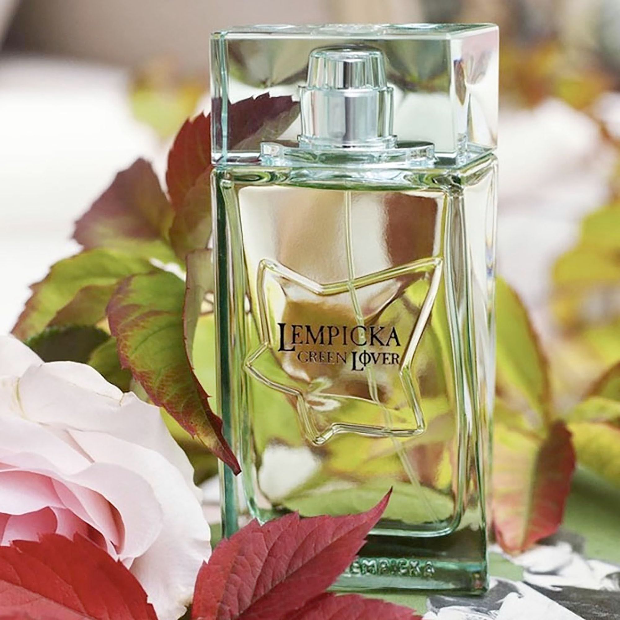 Rechteckiger Flakon mit grünem Deckel. Aufschrift: LEMPICKA GREEN LOVER. Sternförmiges Design auf dem Flakon. Umgeben von Blättern und Blüten.