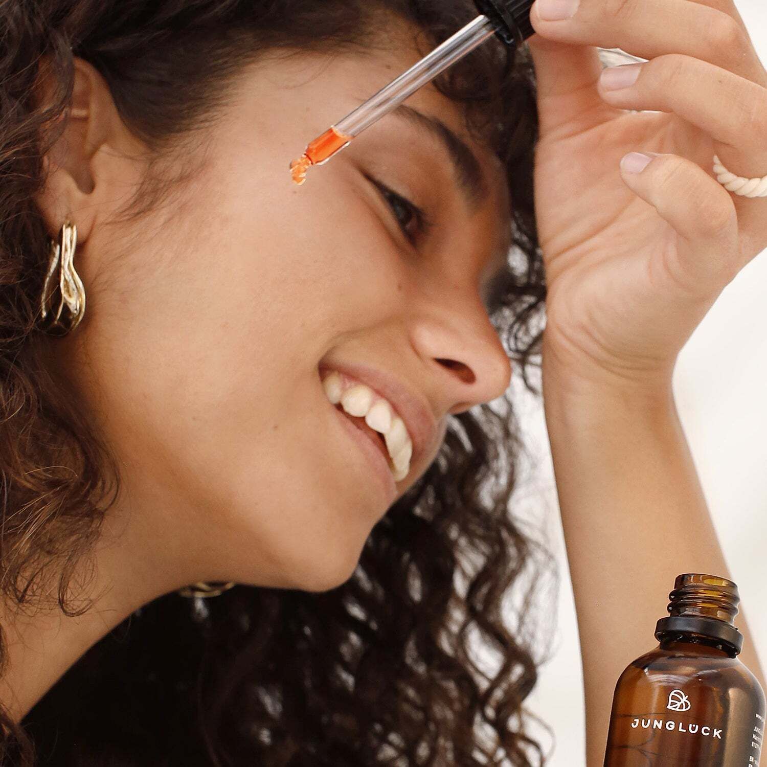 Frau mit Locken hält Pipette mit orangefarbenem Öl. Im Hintergrund: Flasche mit Aufschrift JUNGLÜCK Hagebuttenkernöl.