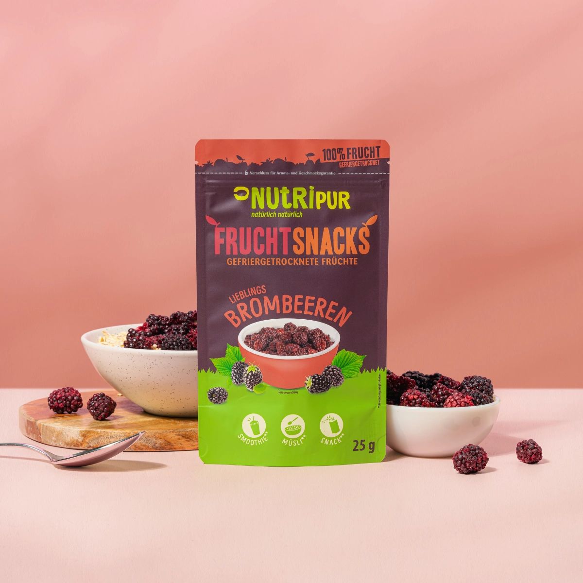 Packung NutriPur Frucht Snacks mit gefriergetrockneten Brombeeren. Daneben Schalen mit Brombeeren und Müsli. Hintergrund rosa.