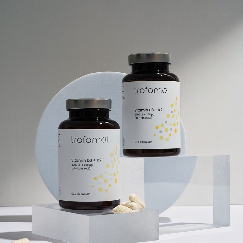 Zwei braune Glasflaschen mit weißem Etikett. TROFOMOL, Vitamin D3 + K2, 120 Kapseln. Daneben Kapseln und weiße Elemente. Silberne Deckel.