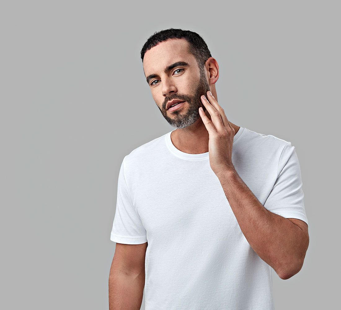 Ein Mann mit Bart berührt sein Gesicht. Er trägt ein weißes T-Shirt.