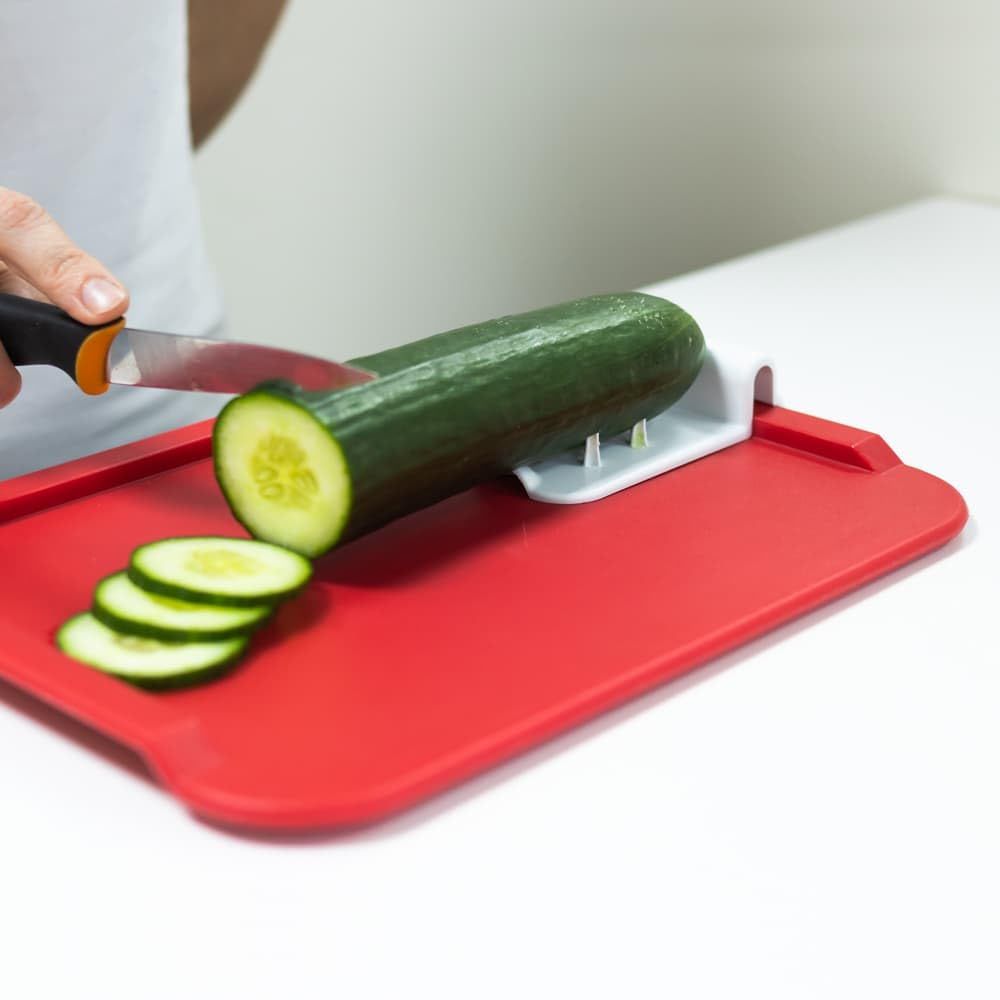 Rotes Essbrettchen. Eine Person schneidet eine Gurke. Die Gurke liegt auf dem Brettchen. Rechteckige Form mit abgerundeten Ecken.
