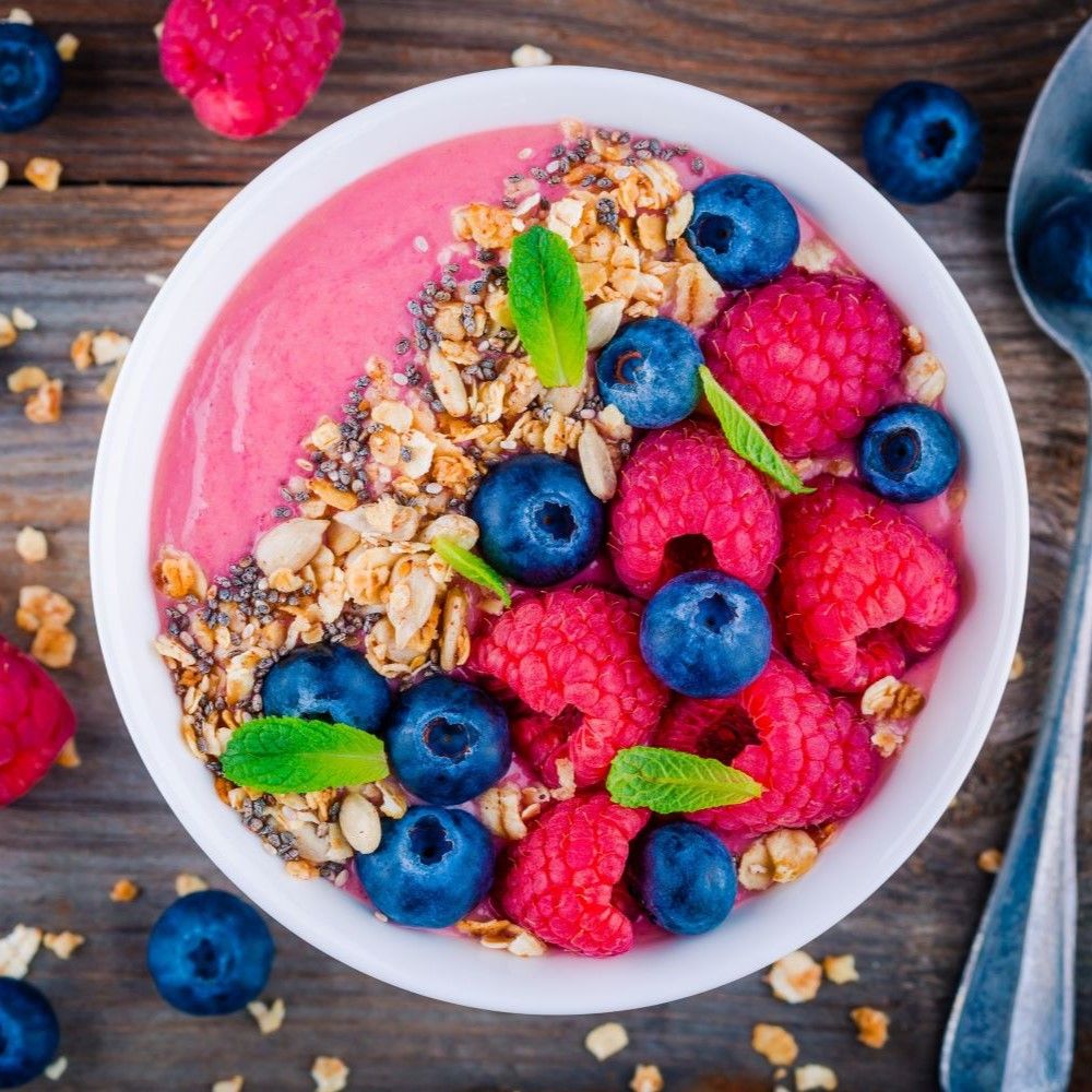 Weiße Schüssel mit pinkfarbenem Smoothie, Beeren und Müsli. Blaubeeren, Himbeeren und Minzblätter.