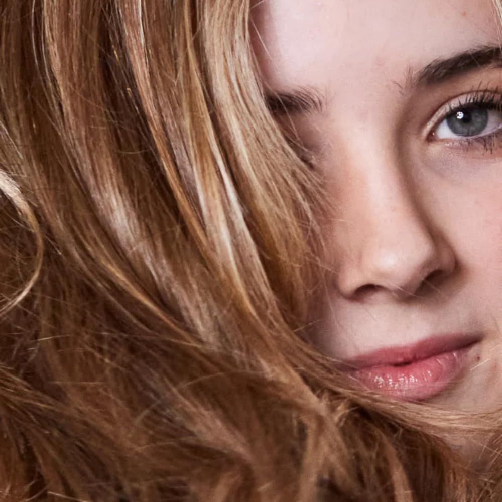 Porträt einer Frau mit welligem, hellbraunem Haar. Ein Teil des Gesichts ist sichtbar, einschließlich Auge und Nase.