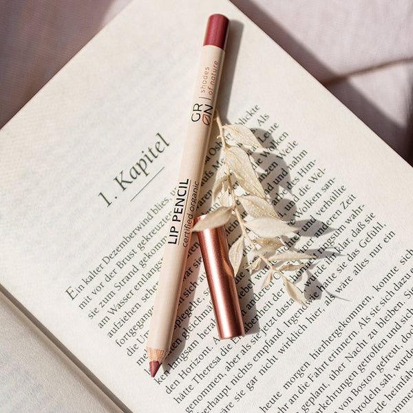 Lippenstiftstift und Kappe liegen auf einem offenen Buch. Aufschrift: LIP PENCIL, GRN [GRÜN].