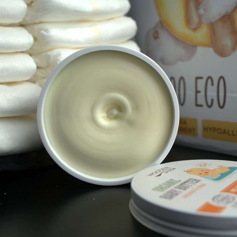 Geöffnete Dose Babybutter. Sichtbar ist die cremige Textur. Im Hintergrund Windeln und Verpackung.