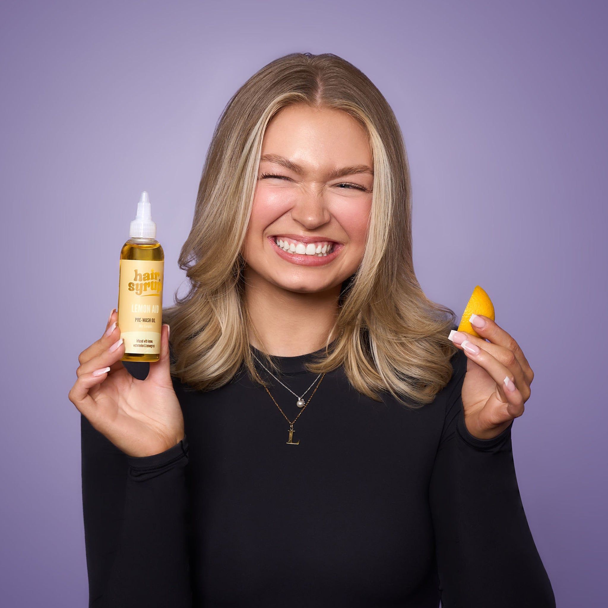 Frau hält eine Flasche "Hair Syrup Vitamin C Me" und eine Orangenscheibe. Pre-Wash Oil. Lila Hintergrund.