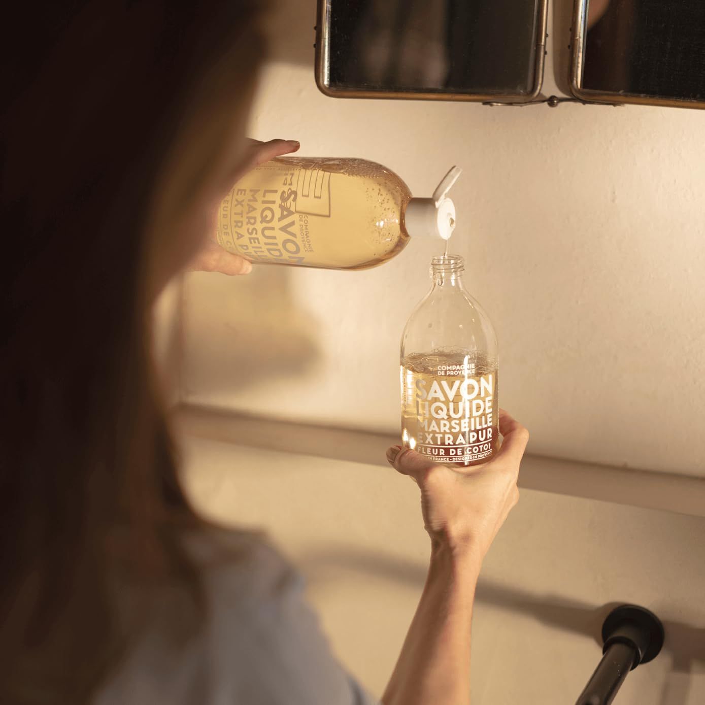 Person gießt Flüssigseife aus einer Flasche in eine andere. Beide Flaschen sind durchsichtig und beschriftet.