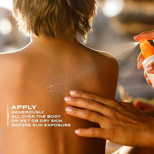 Person sprüht Sonnencreme auf den Rücken. Weiße Flasche mit orangefarbenem Sprühkopf. Aufschrift: Lancaster.