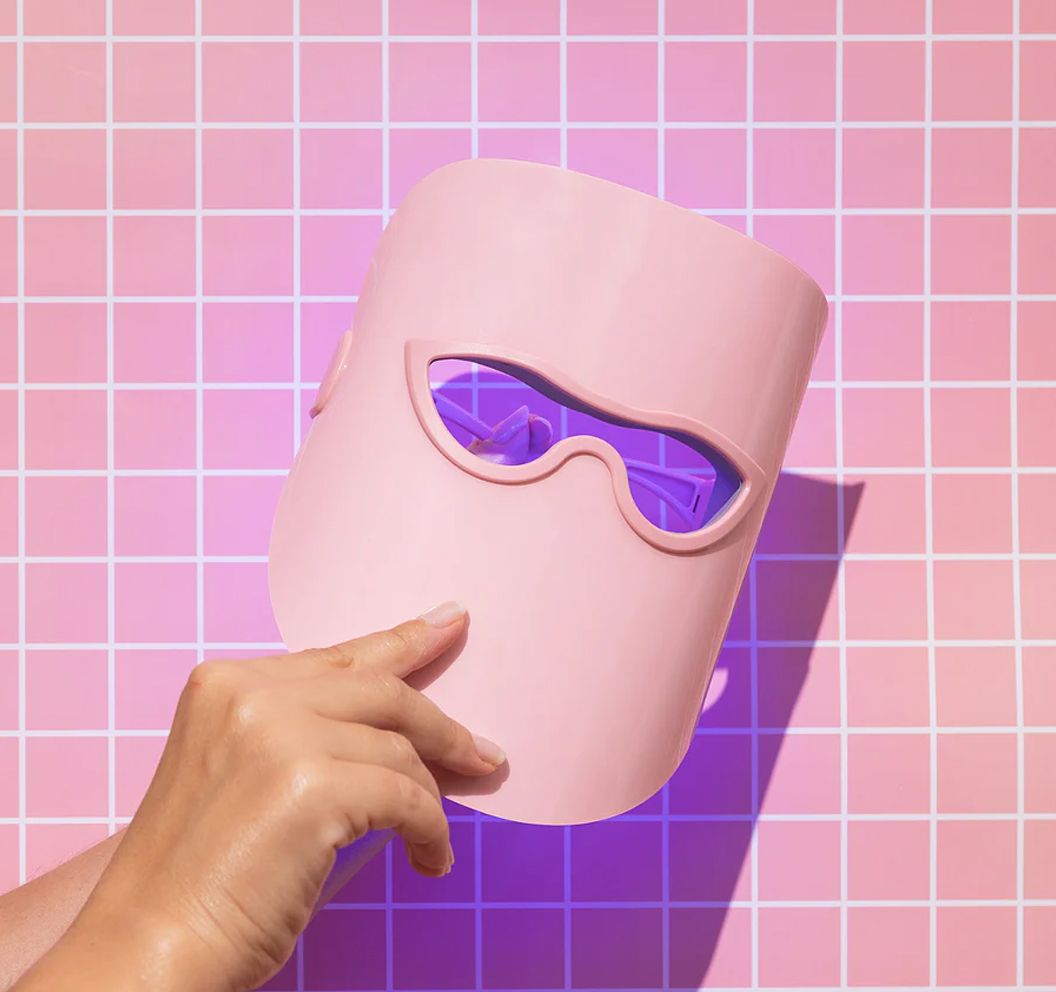 Eine rosafarbene LED-Maske wird in einer Hand vor einem karierten Hintergrund gehalten.