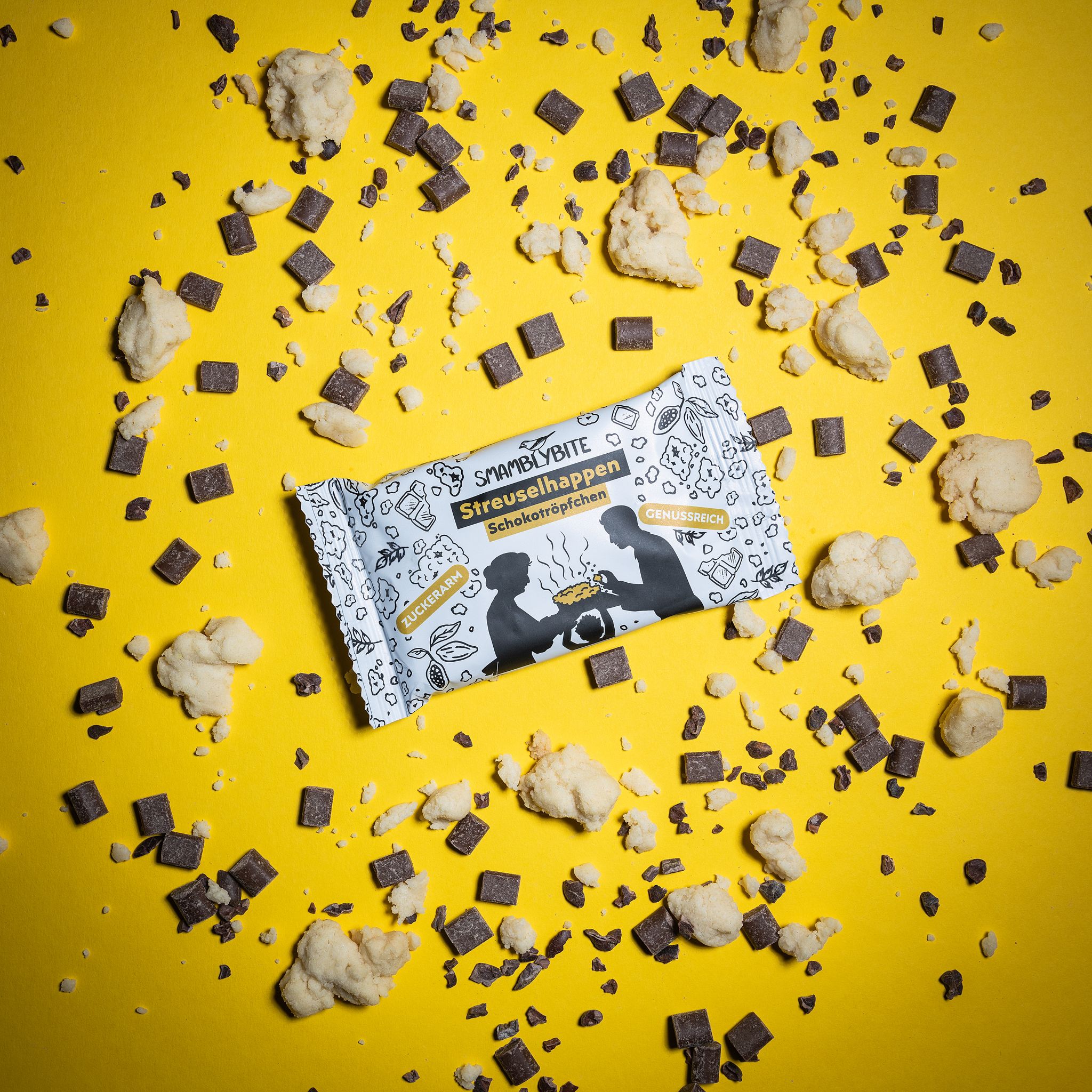 SMAMBLYBITE Streuselhappen-Riegel auf gelbem Hintergrund, umgeben von Streuseln und Schokostückchen. Verpackung mit Logo und Text.