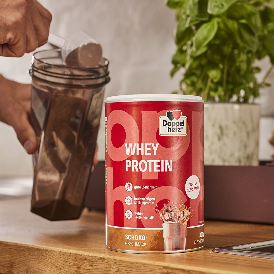 Person gießt Proteinpulver in einen Shaker. Dose Doppelherz WHEY PROTEIN Schoko im Hintergrund.