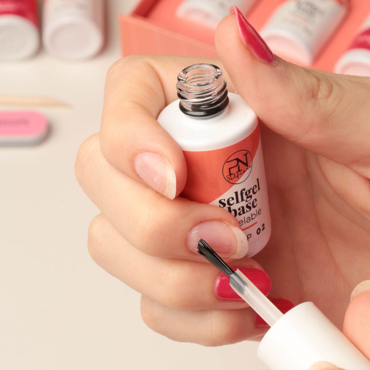 Hand hält Flasche mit Pinsel. Text: selfgel base peelable, STEP 02. PN-Logo. Nagel mit rotem Nagellack.