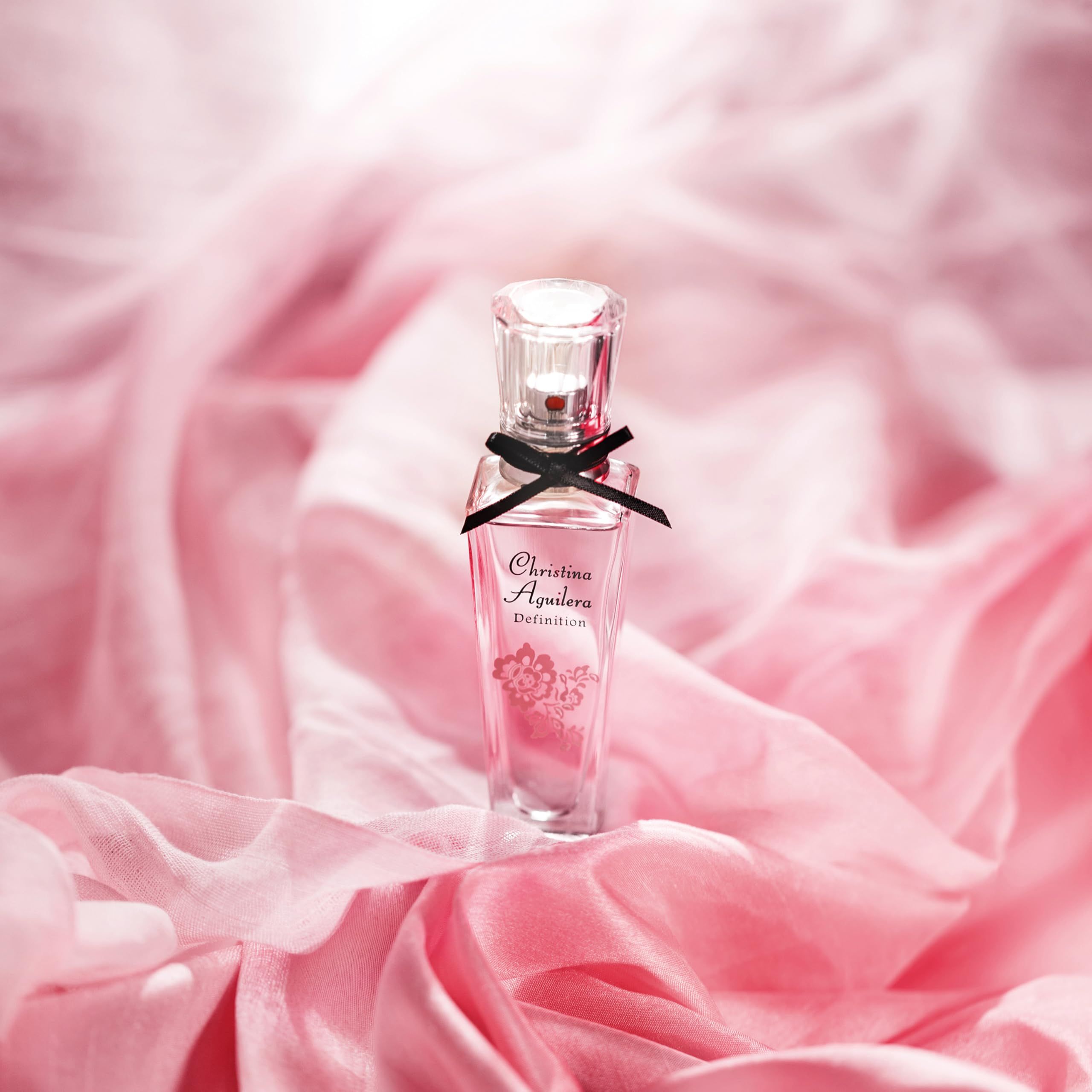 Parfümflakon mit rosa Flüssigkeit, schwarzer Schleife und floralem Design. Aufschrift: Christina Aguilera Definition. Hintergrund: Stoff.