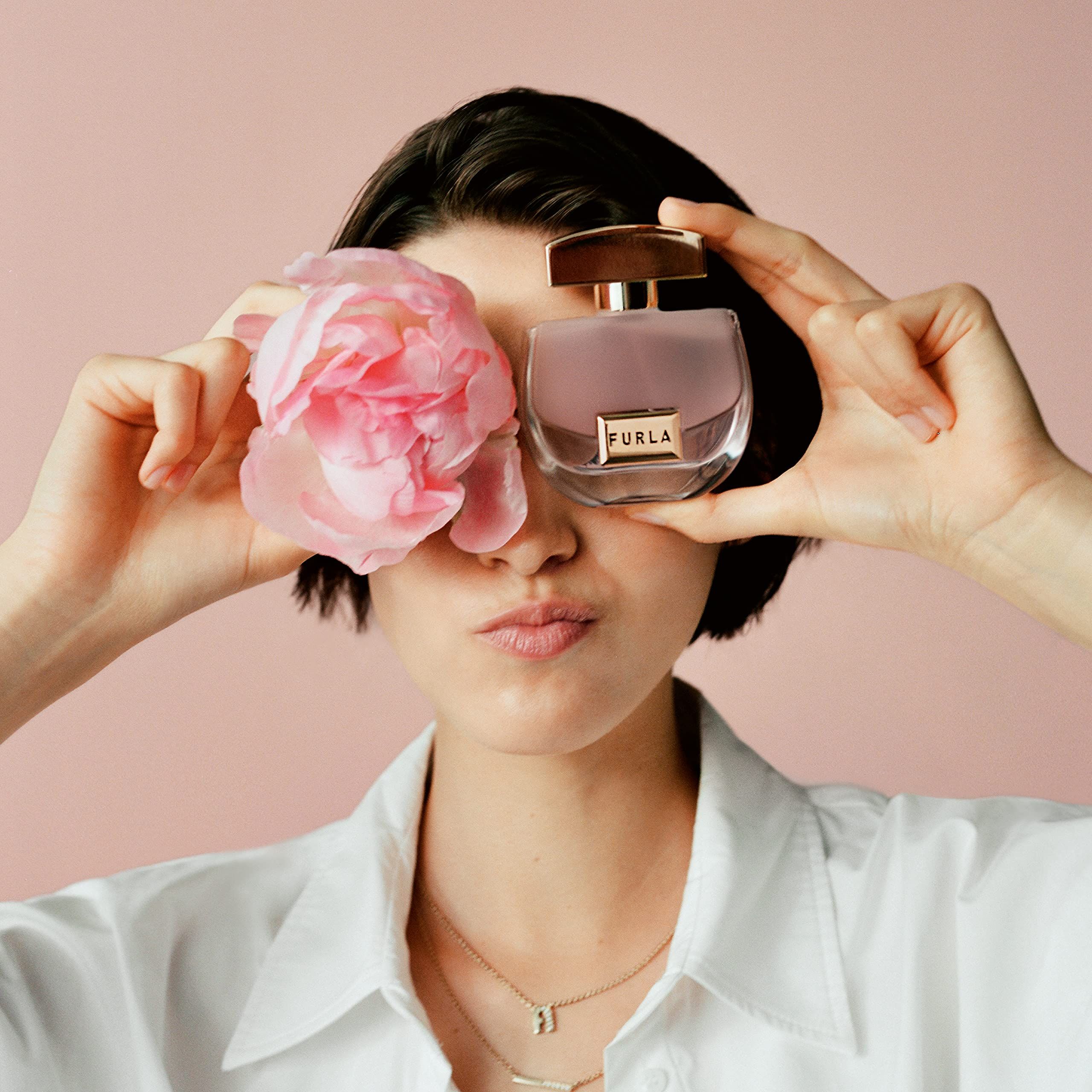 Frau hält Parfümflakon und Blume vor Augen. Flakon mit goldenem Deckel und FURLA-Logo.