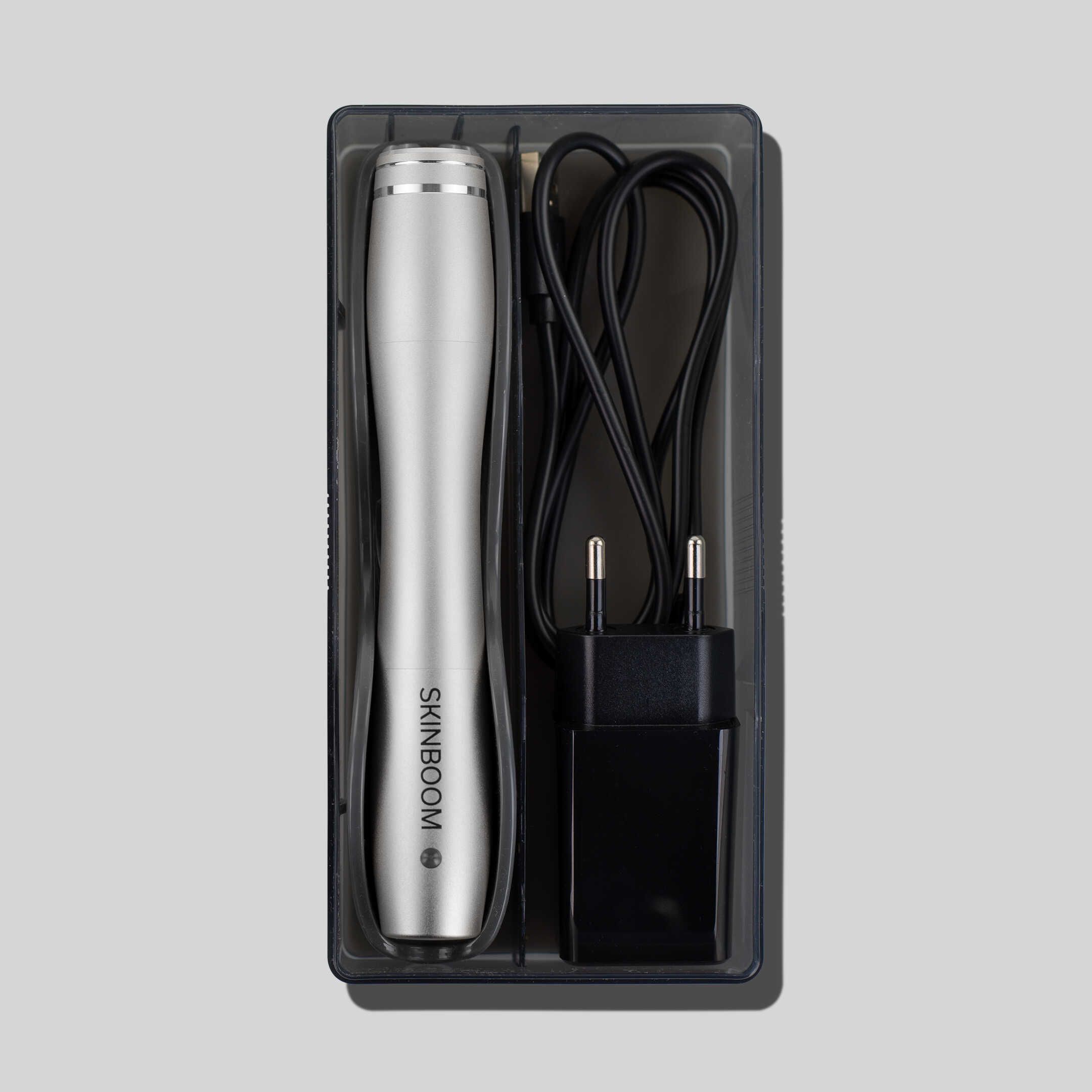 SKINBOOM-Gerät in einer schwarzen Box mit Netzteil und Ladekabel. Das Gerät ist silberfarben.