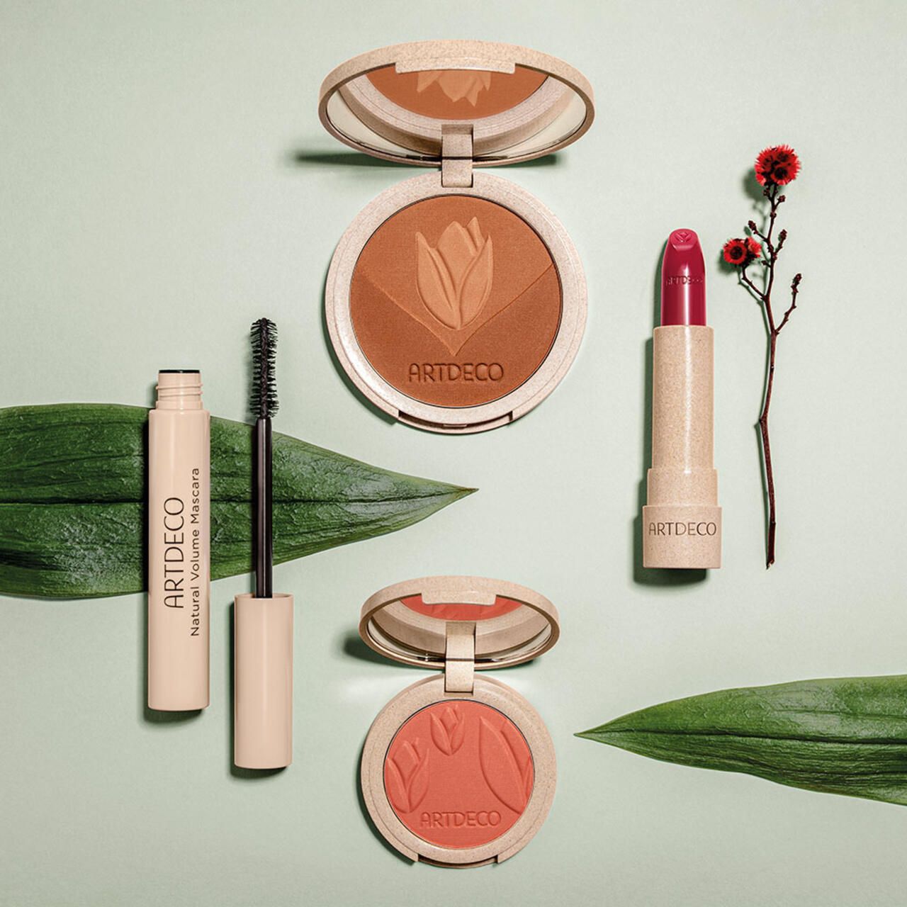 Verschiedene Artdeco-Kosmetikprodukte: Bronzer, Mascara, Lippenstift. Auf grünem Hintergrund.