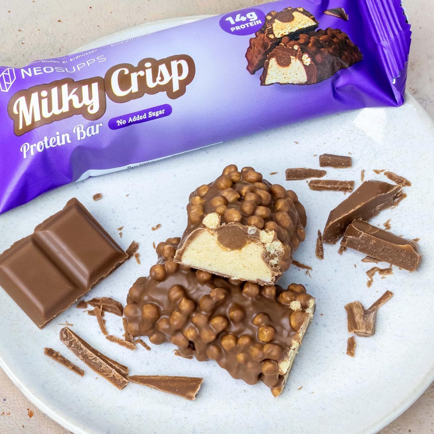 Ein Proteinriegel, halbiert, auf einem Teller. Schokoladenstücke und weitere Riegelteile daneben. Verpackung im Hintergrund.