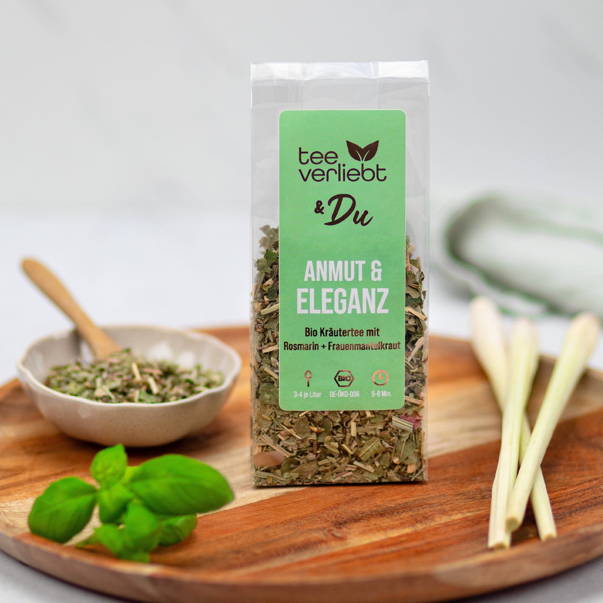 Transparente Verpackung mit grünem Etikett. Aufschrift: teeverliebt & Du, Anmut & Eleganz. Kräutertee auf Holzbrett mit Kräutern.