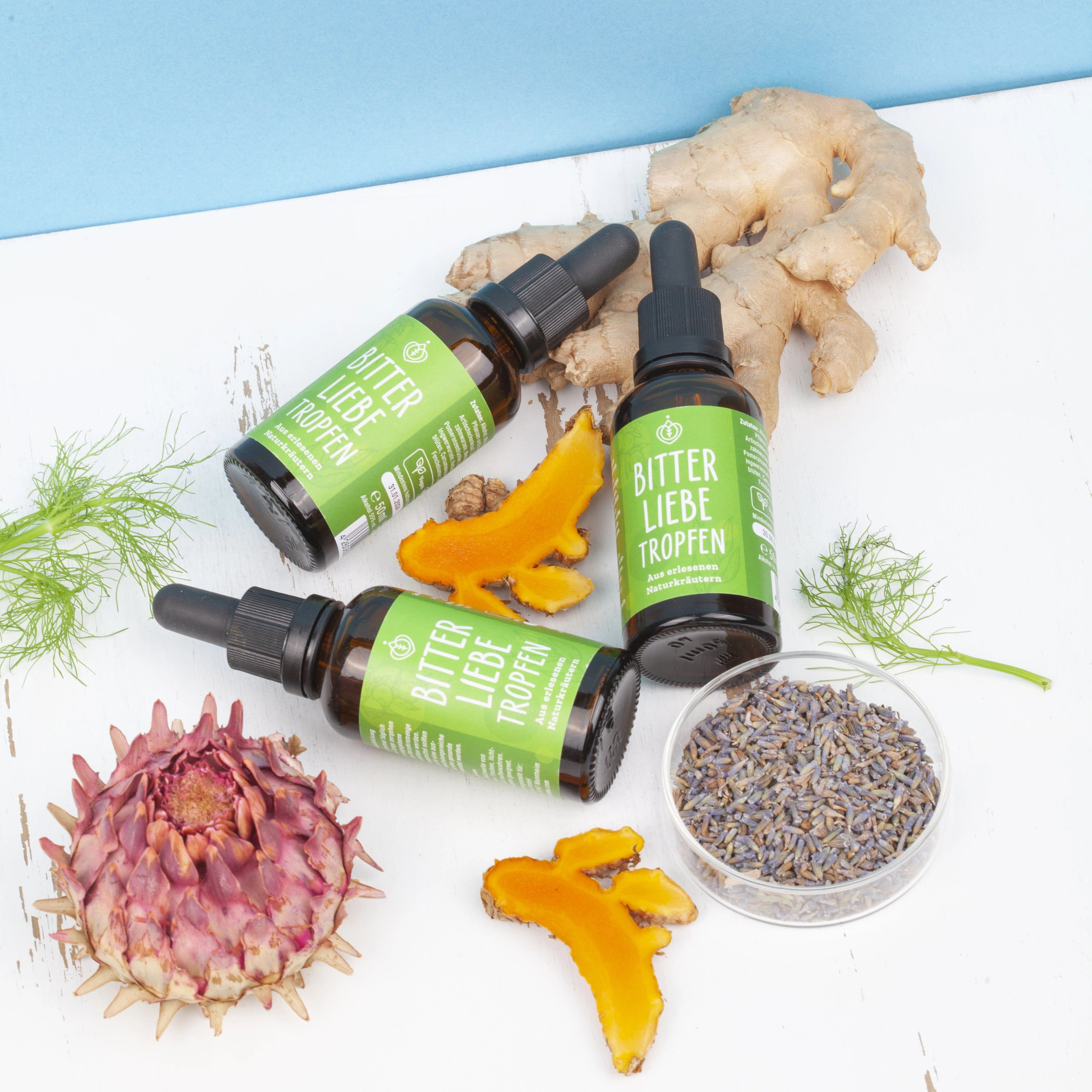 Drei Flaschen BitterLiebe Tropfen. Braune Flaschen mit grüner Beschriftung. Kräuter und Ingwer daneben.