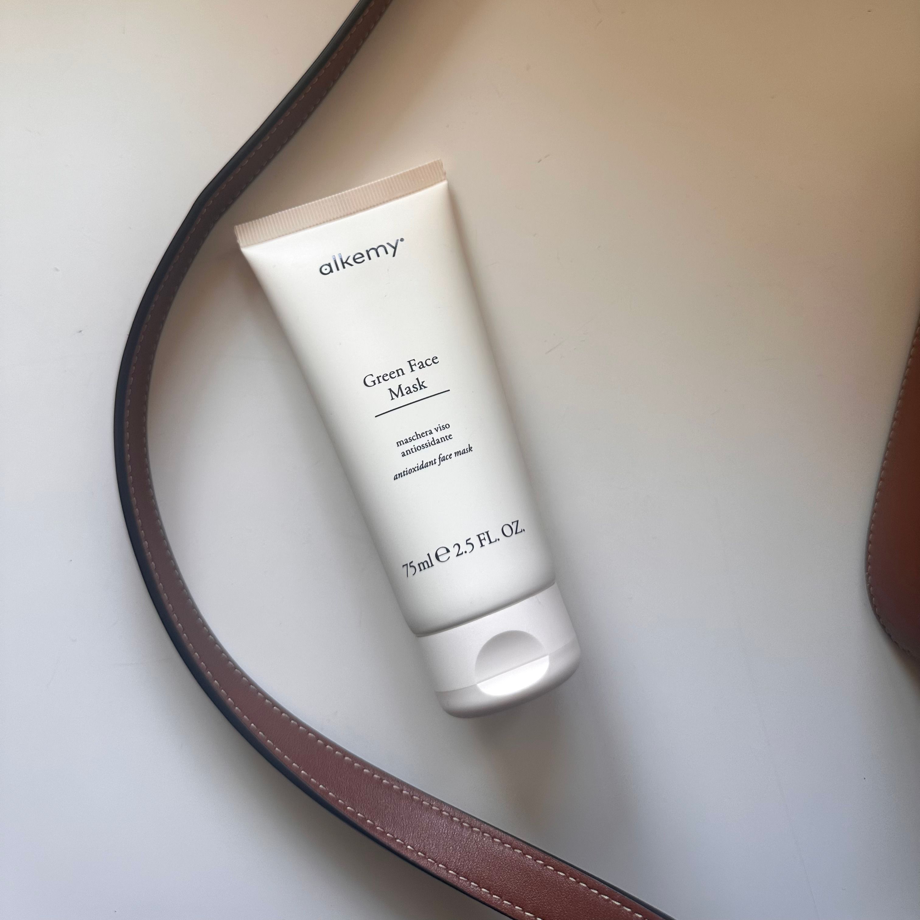 Weiße Tube Alkemy Green Face Mask neben einer braunen Ledertasche.