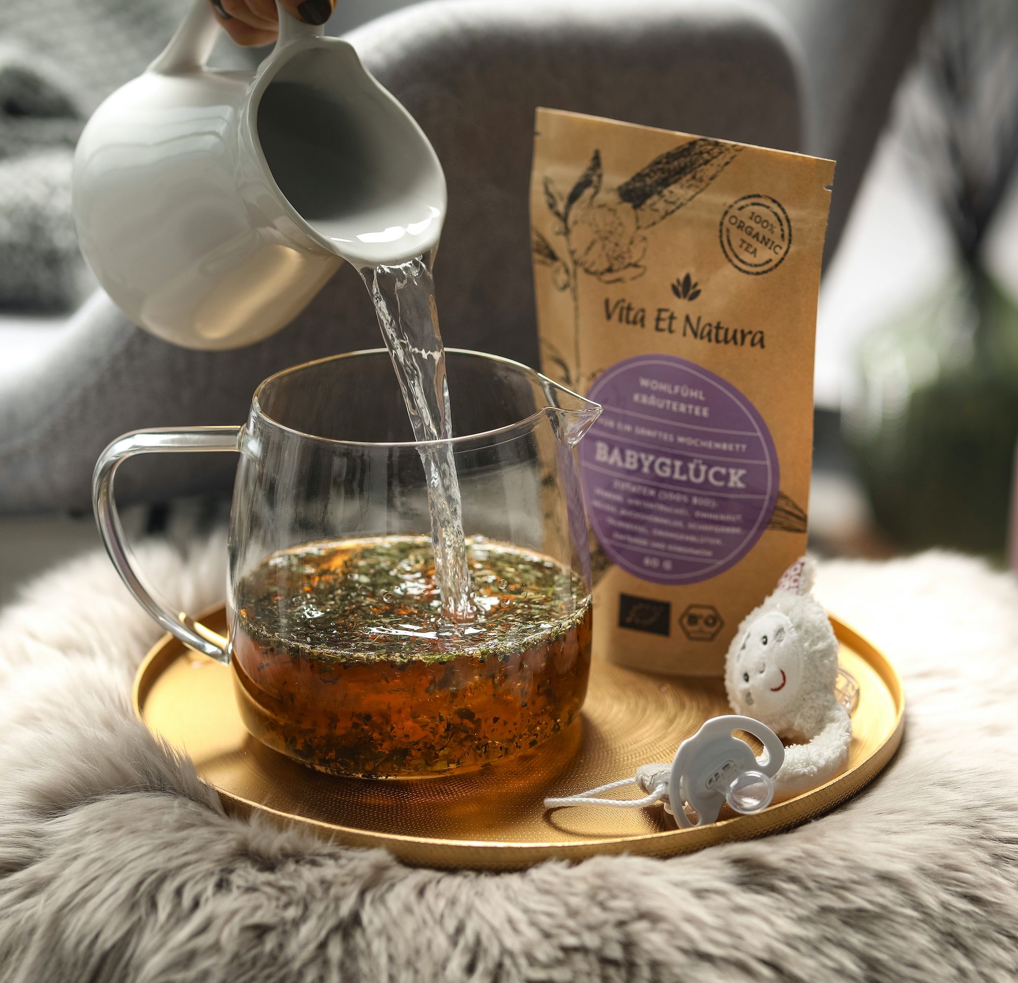 Teezubereitung. Kanne mit heißem Wasser, Teepackung Babyglück, Tablett mit Tasse. Kuscheltier.