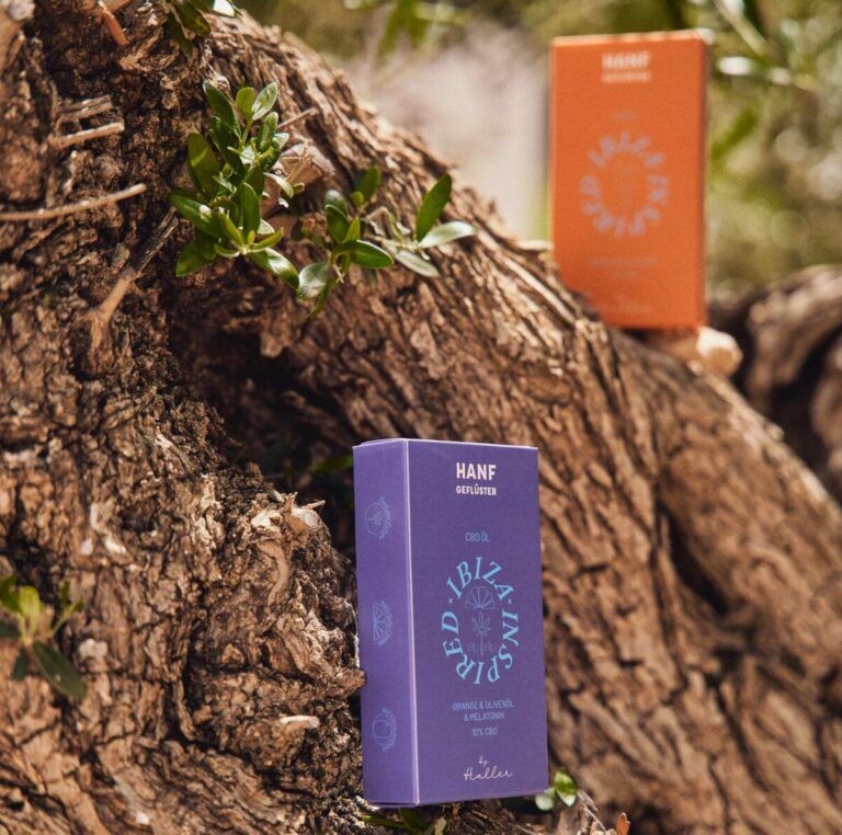 Zwei Schachteln, lila und orange, mit Text: HANF, CBD, Ibiza Spirit. Auf einem Baumstamm. Von Haller.