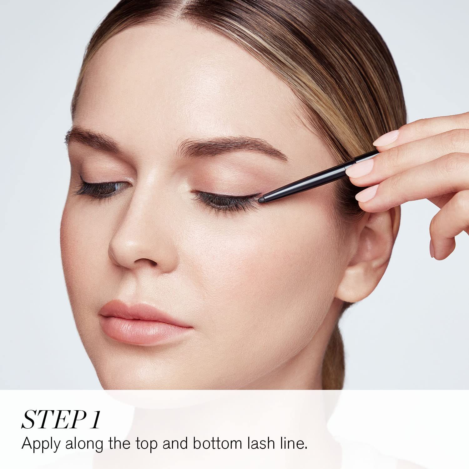 Nahaufnahme einer Frau, die einen schwarzen Eyeliner auf das obere Augenlid aufträgt. Text: Schritt 1. Auf den oberen und unteren Wimpernkranz auftragen.