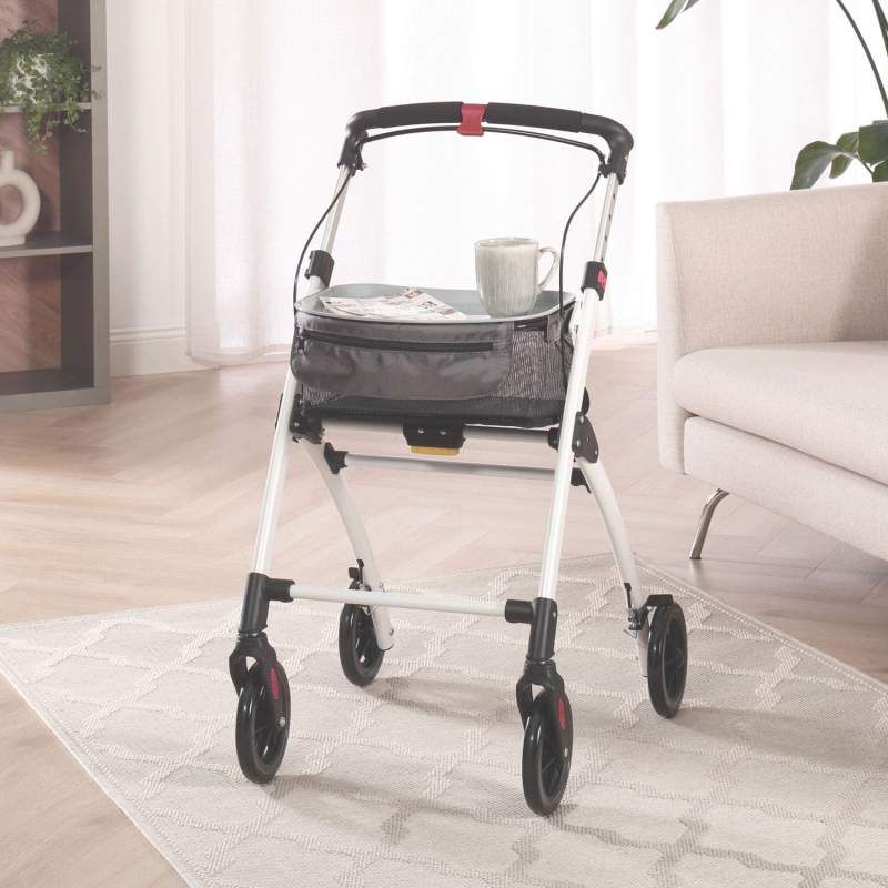 Rollator im Wohnzimmer. Weißer Rahmen, schwarze Räder und Korb. Graue Tasche. Auf dem Tablett: Tasse und Teller.
